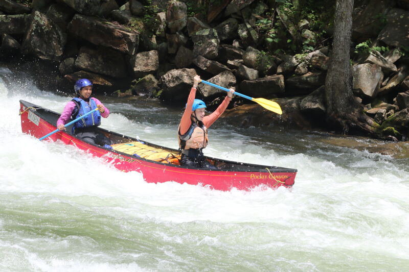 Two people are navigating a red canoe through whitewater rapids. The person in the front is paddling, while the person in the back is holding their paddle up. They are both wearing helmets and life jackets. The water is turbulent and foamy, and the background shows rocks and trees.